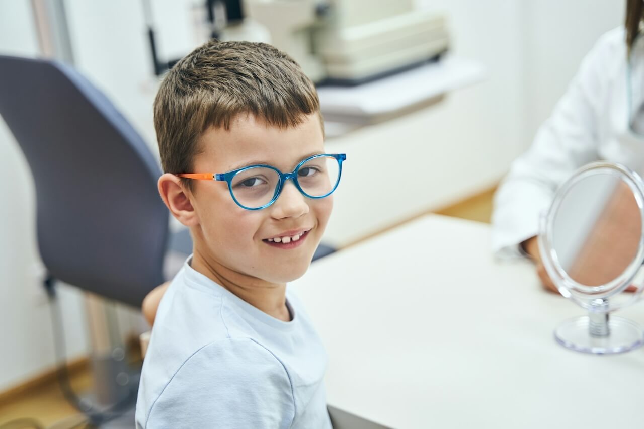 En la fotografía se aprecia un niño sonriente con gafas azules sentado en una consulta oftalmológica, simbolizando la importancia del control de la vista en la infancia.